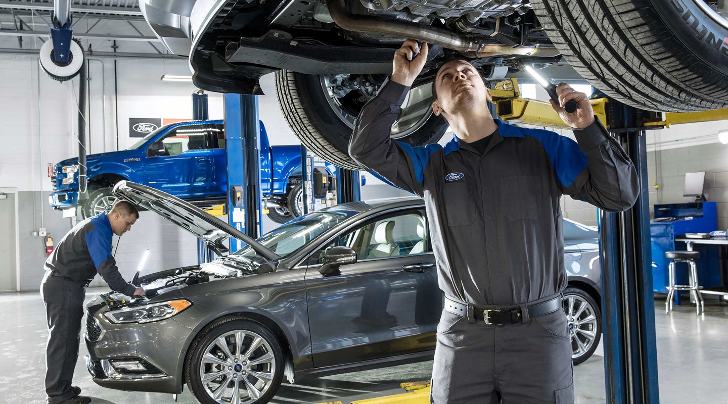 A Certified Ford Service Technician performing maintenance beneath a Ford vehicle, with another Certified Ford Service Technician performing maintenance under the hood of another Ford vehicle in the background
