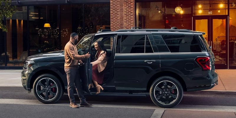 A man helping a woman out of the drivers side of a dark teal blue 2024 Ford Expedition Max that is parked on a city street at dusk