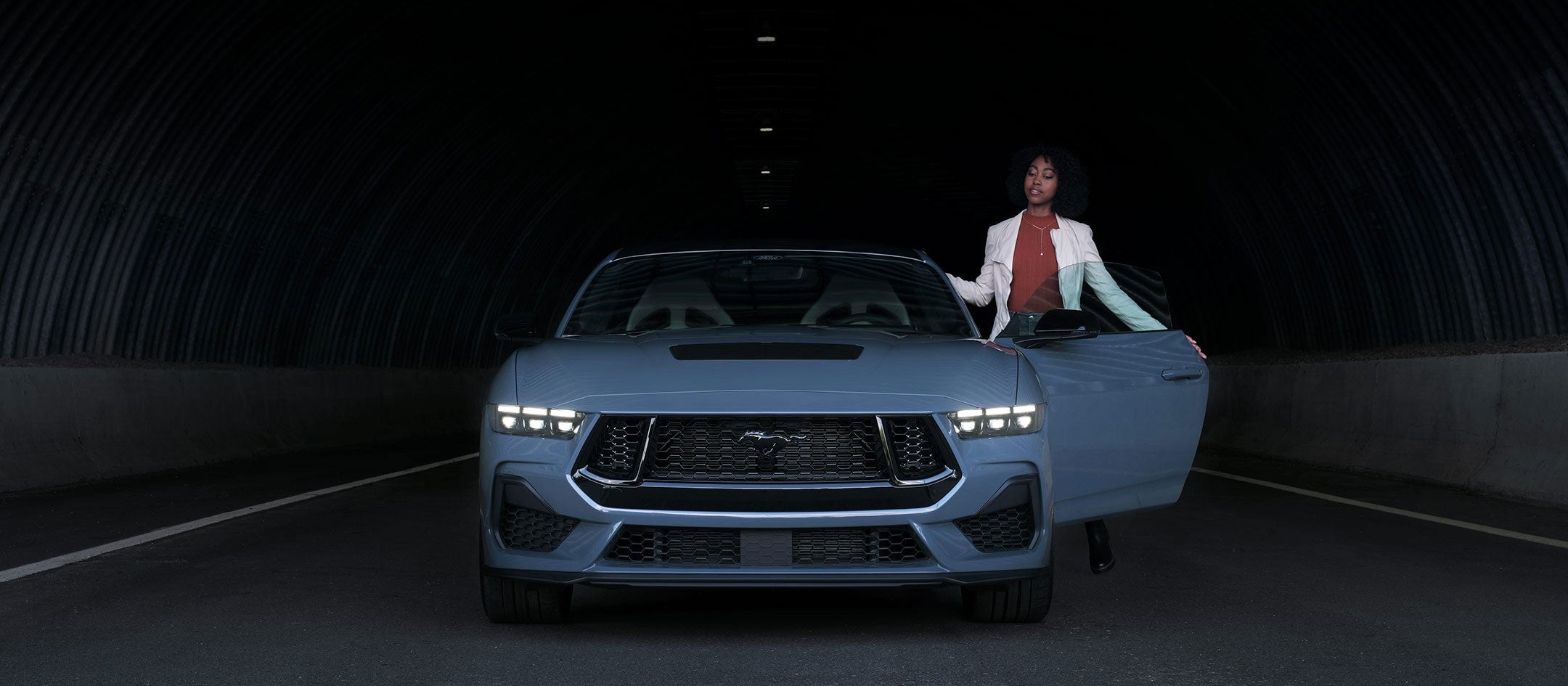 A woman standing and holding open the drivers side door of a light blue 2024 Ford Mustang
