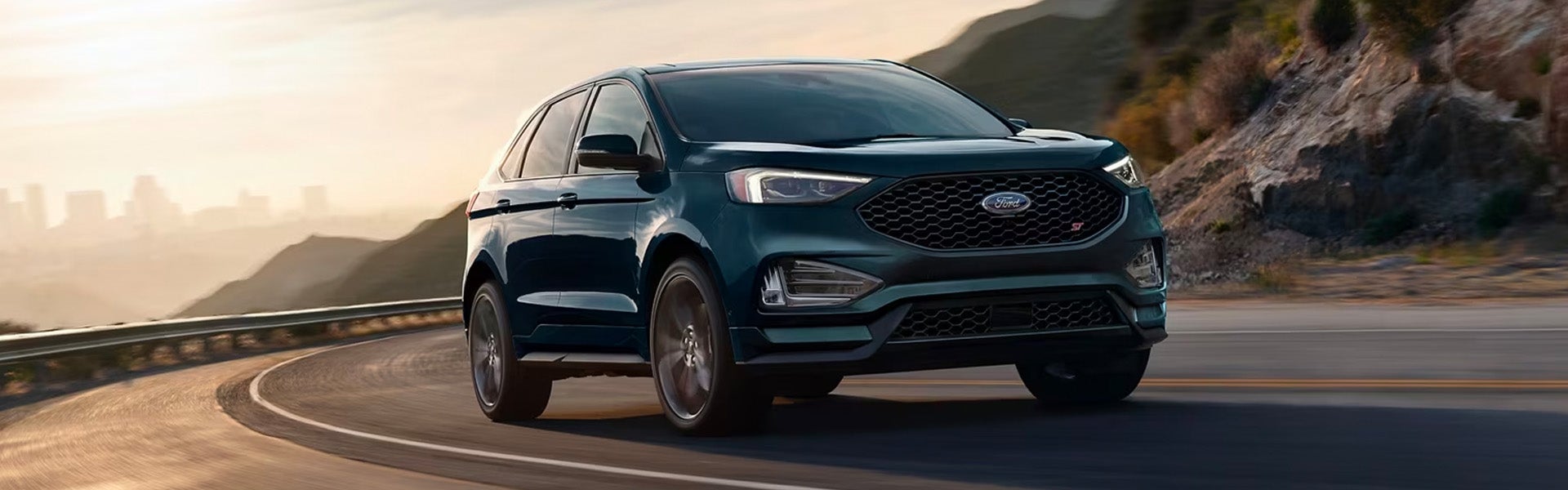 A teal blue 2024 Ford Edge driving down a coastal highway when the sun is setting